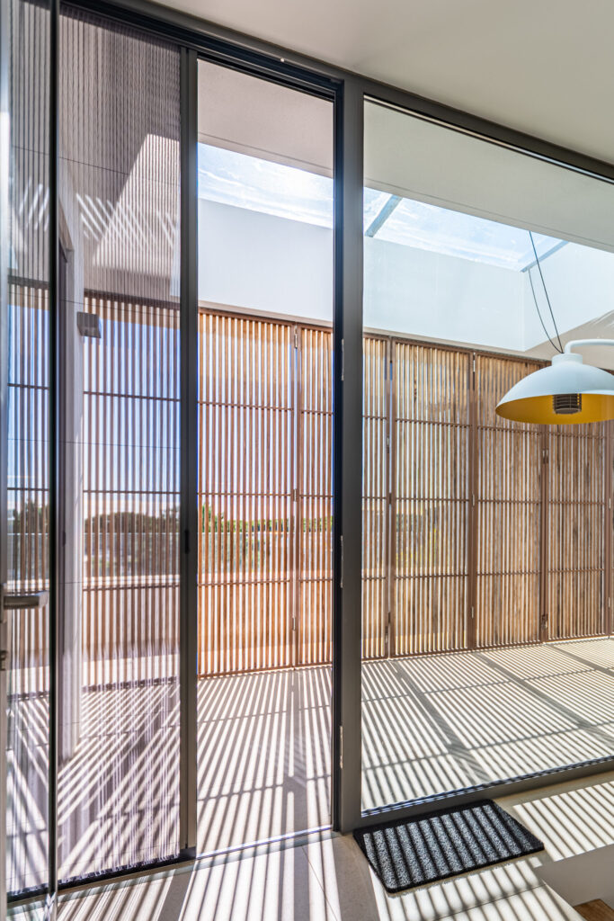 Contemporary corridor with a pleated plisse screen on sliding doors, showcasing sunlight patterns and wooden outdoor panels.