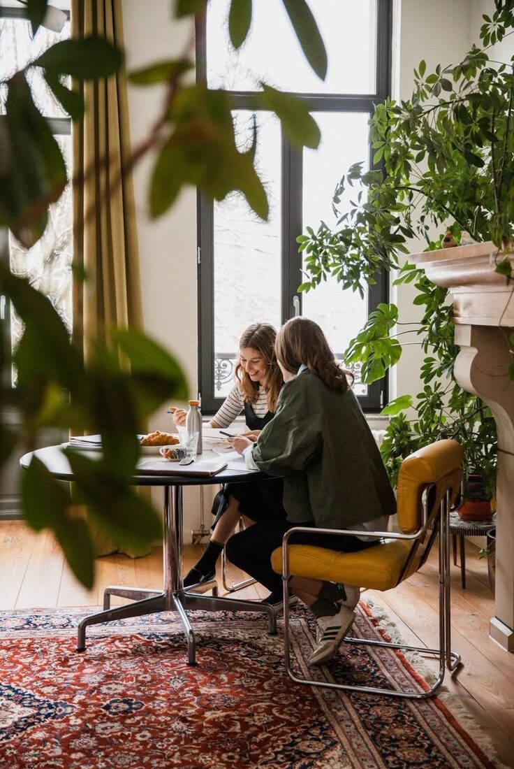 Two women sitting at a table working in a bright room filled with plants and natural decor.